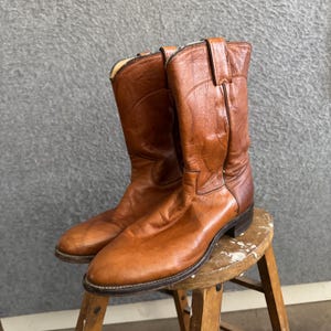May include: A pair of mid-calf western-style boots crafted from smooth, tan-coloured leather. These boots feature a rounded toe, a low stacked heel, and vertical pull tabs at the top of the shaft, showcasing a classic design with visible stitching.
