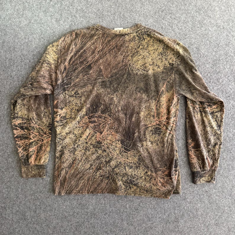 May include: Long-sleeved shirt with a camouflage pattern in shades of brown, tan, and green. The shirt has a crew neck and is made of a soft, possibly cotton, material. The camouflage design is detailed and covers the entire shirt.