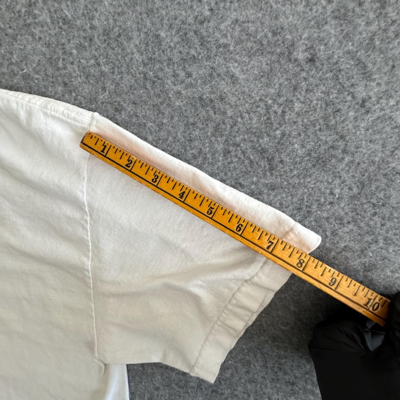 May include: A white t-shirt sleeve is measured with a yellow ruler. The ruler shows measurements up to 10 inches. The t-shirt is on a gray surface.