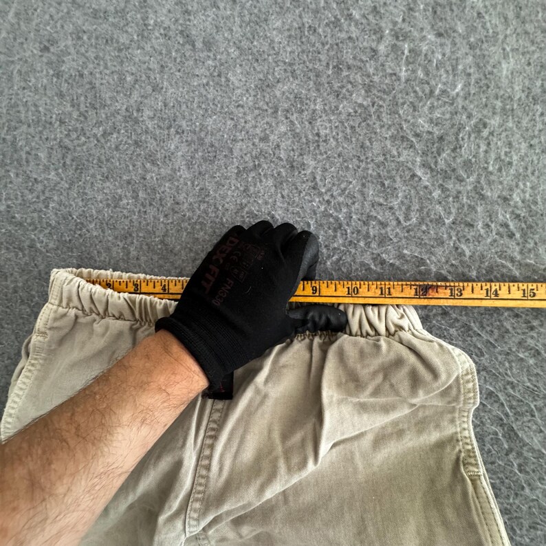K&ouml;nnte beinhalten: Eine beigefarbene Hose mit elastischem Bund wird mit einem gelben Ma&szlig;band gezeigt, das oben angelegt ist und eine Bundweite von etwa 38 cm von der linken bis zur rechten Kante des Kleidungsst&uuml;cks anzeigt.
