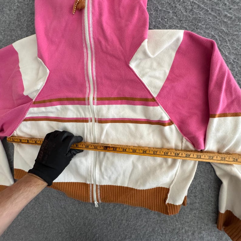Op de afbeelding: Een roze, witte en bruine hoodie met ritssluiting. De sweatshirt heeft een witte rits, bruine geribde manchetten en een wit en bruin gestreept ontwerp over de borst. De borstbreedte is ongeveer 61 cm.