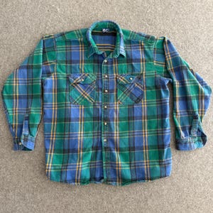 Op de afbeelding: Een groen, blauw en geel geruit flanellen overhemd. Het shirt met lange mouwen heeft twee borstzakken en een knoopsluiting aan de voorkant. De kraag is een donkerdere tint groen. Het shirt is gemaakt van zachte stof.