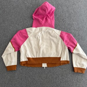 Op de afbeelding: Korte hoodie in cr&egrave;me, roze en bruin. De achterkant van de jas heeft een roze capuchon, roze schouderpanelen en bruine geribde manchetten en zoom. Een bruine streep loopt over de onderkant en een rits is zichtbaar.