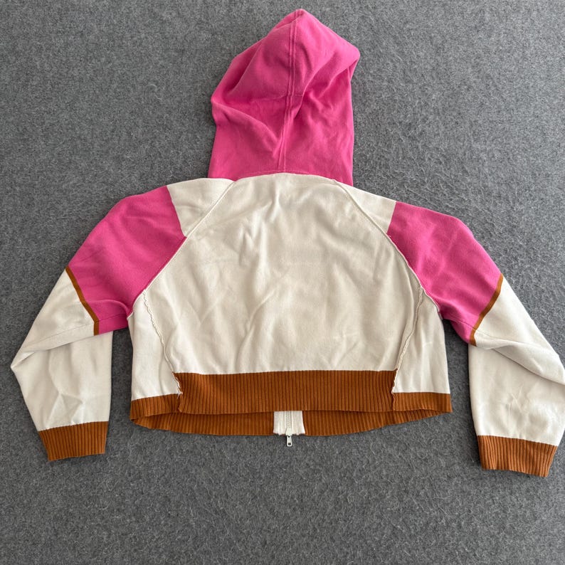 Op de afbeelding: Korte hoodie in cr&egrave;me, roze en bruin. De achterkant van de jas heeft een roze capuchon, roze schouderpanelen en bruine geribde manchetten en zoom. Een bruine streep loopt over de onderkant en een rits is zichtbaar.