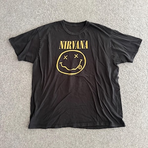 May include: A black short-sleeve t-shirt featuring the word NIRVANA printed in yellow capital letters above a yellow graphic of a smiley face with X-shaped eyes and a tongue sticking out from the mouth, centered on the chest area.