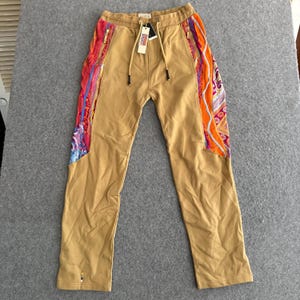 May include: Tan sweatpants featuring a drawstring waist and side panels with a vibrant, multi-colored patterned knit design. The pants include gold-toned zippers on the side pockets and a white brand tag attached to the front drawstring.