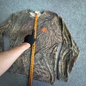 May include: Long-sleeved camouflage shirt with a crew neck. The shirt features a brown, green, and tan pattern, with an orange logo on the chest. The shirt is being measured with a wooden ruler.