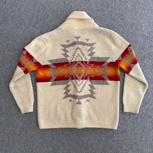 May include: A cream-colored knit cardigan featuring a large, centered geometric pattern on the back. The design incorporates horizontal bands of red, orange, and grey, extending across the shoulders and down the sleeves with ribbed cuffs.