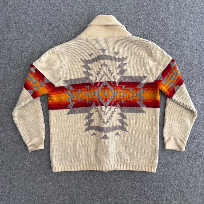 May include: A cream-colored knit cardigan featuring a large, centered geometric pattern on the back. The design incorporates horizontal bands of red, orange, and grey, extending across the shoulders and down the sleeves with ribbed cuffs.