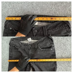 Pu&ograve; includere: Jeans in denim nero con chiusura a bottone, passanti e tasche. Un righello mostra che la misura della vita &egrave; di circa 40,6 cm. I jeans sono appoggiati su una superficie grigia.