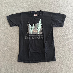Peut inclure: Un t-shirt noir à manches courtes orné d'un motif de sapins stylisés et d'une structure centrale. Sous le motif, le texte indique : Celebrating a Season of Fresh New Snow, Nutcracker, Berkeley Ballet Theater en lettres blanches.