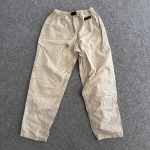 K&ouml;nnte beinhalten: Beigefarbene Hose mit elastischem Bund und schwarzem Kordelzug. Die Hose hat ein kleines rechteckiges schwarzes Etikett mit rotem Text auf der oberen rechten Seite und einen sichtbaren Riss im Stoff im Bereich der mittleren Schrittnaht.