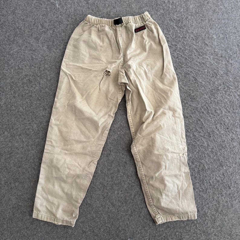 K&ouml;nnte beinhalten: Beigefarbene Hose mit elastischem Bund und schwarzem Kordelzug. Die Hose hat ein kleines rechteckiges schwarzes Etikett mit rotem Text auf der oberen rechten Seite und einen sichtbaren Riss im Stoff im Bereich der mittleren Schrittnaht.