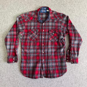 Könnte beinhalten: Rot-grau-blau kariertes Button-up-Hemd. Das langärmlige Hemd hat zwei Brusttaschen und einen blauen Kragen. Das Hemd ist aus einem weichen, gewebten Stoff gefertigt. Der Markenname ist am Kragen sichtbar.