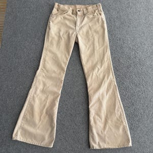 May include: A pair of beige corduroy pants featuring a high-waisted design, a button and zipper fly closure, two front pockets, and a flared leg silhouette. The fabric has a distinct vertical ribbed texture throughout the entire garment.