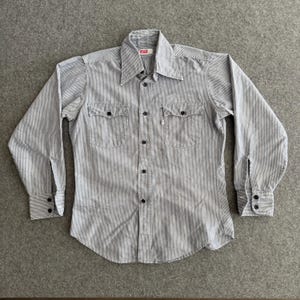 Puede incluir: Camisa a rayas verticales azules y blancas, de manga larga y abotonada. Tiene dos bolsillos en el pecho, botones negros y cuello. El logotipo de Levi's es visible en la etiqueta.