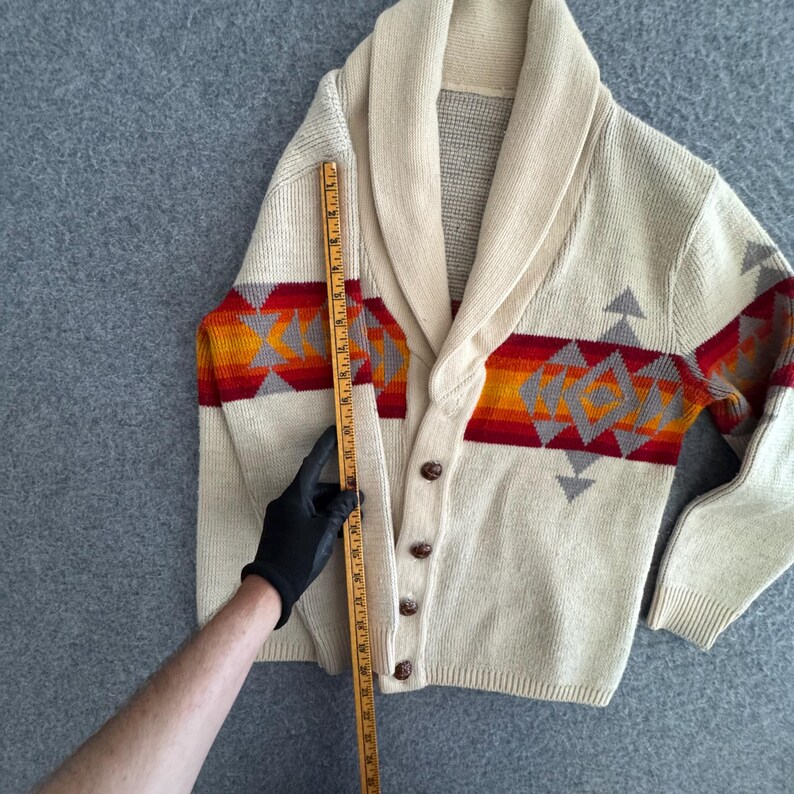 May include: A cream-colored knit cardigan with a shawl collar and a horizontal band featuring a geometric pattern in red, orange, and gray. A wooden ruler is placed vertically along the front, measuring approximately 27 inches in length.
