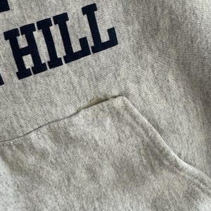 May include: Close-up of a light gray hoodie with a front pocket. The word "HILL" is printed in navy blue block letters across the top. The fabric shows signs of wear and tear, with a small hole near the pocket.