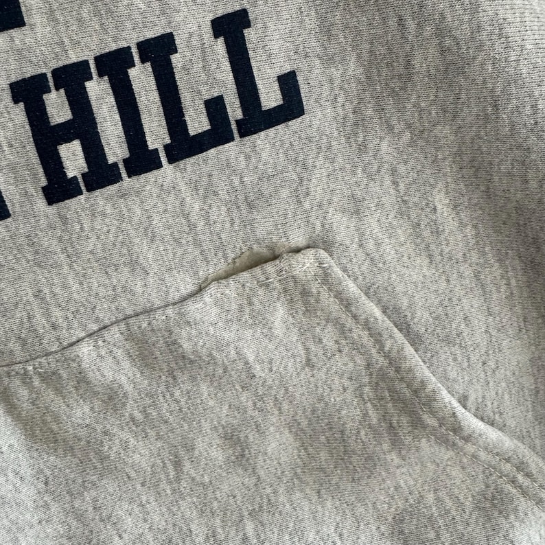 May include: Close-up of a light gray hoodie with a front pocket. The word "HILL" is printed in navy blue block letters across the top. The fabric shows signs of wear and tear, with a small hole near the pocket.