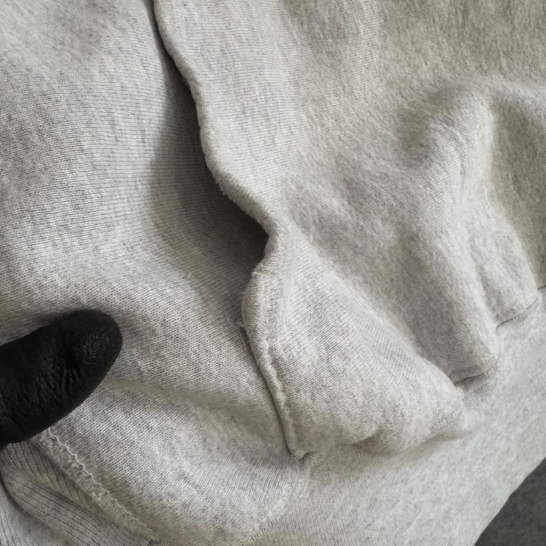 May include: Close-up of a light gray sweatshirt pocket. The fabric is a soft, textured material. The pocket is sewn into the garment with visible stitching. A black object is partially visible in the lower left corner.