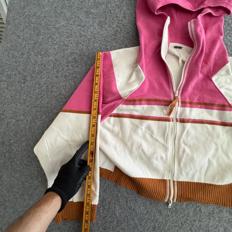 Op de afbeelding: Een roze, witte en bruine hoodie met ritssluiting. De sweatshirt heeft een witte body met roze en bruine accenten. De capuchon en de bovenmouwen zijn roze. De zoom en manchetten zijn bruin. De lengte wordt gemeten met een liniaal.
