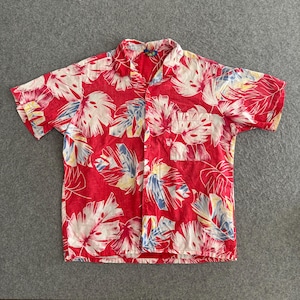 Puede incluir: Camisa roja de manga corta con un estampado de hojas tropicales en blanco, amarillo y azul. La camisa tiene un bolsillo en el pecho y un cuello clásico. La tela parece ligera.