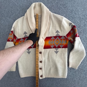 May include: A cream-colored knit cardigan with a shawl collar and four brown buttons. A horizontal band features a geometric pattern in red, orange, and gray. A wooden ruler placed vertically against the center shows a length of 24 inches.