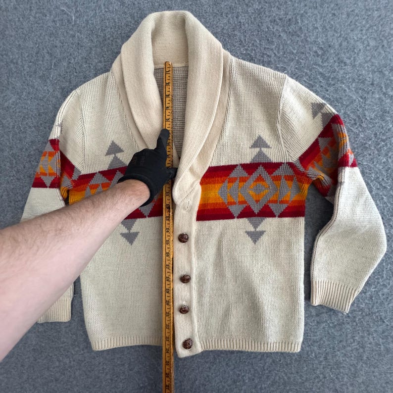 May include: A cream-colored knit cardigan with a shawl collar and four brown buttons. A horizontal band features a geometric pattern in red, orange, and gray. A wooden ruler placed vertically against the center shows a length of 24 inches.
