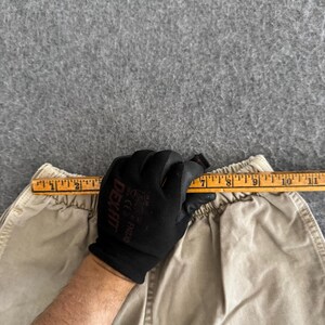 K&ouml;nnte beinhalten: Eine beigefarbene Hose mit elastischem Bund wird mit einem Holzlineal gezeigt, das oben quer liegt. Das Lineal zeigt ein Ma&szlig; von etwa 25,4 cm im Taillenbereich an, wobei die Markierungen auf dem gelben Holz deutlich sichtbar sind.