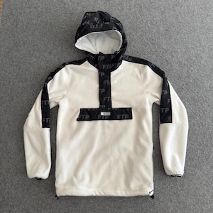 May include: A white fleece hoodie with a black patterned hood and accents. The hood and sleeves feature a repeating pattern of the letters "FTP". The front pocket and upper chest area also display the "FTP" logo. The hoodie has a drawstring and a zipper on the side.