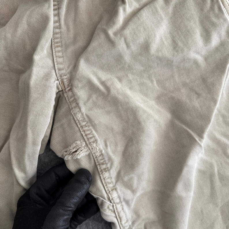 K&ouml;nnte beinhalten: Eine beigefarbene Baumwollhose mit einem sichtbaren Riss entlang der inneren Beinnaht. Der Stoff weist eine ausgepr&auml;gte K&ouml;perbindung auf und das Kleidungsst&uuml;ck ist mit verst&auml;rkten Doppeln&auml;hten im gesamten Beinbereich verarbeitet.