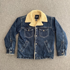 Puede incluir: Chaqueta vaquera azul con cuello y forro de borreguillo beige. La chaqueta tiene cierres de botón, dos bolsillos en el pecho y dos bolsillos laterales. La marca "GAP" es visible en el cuello.