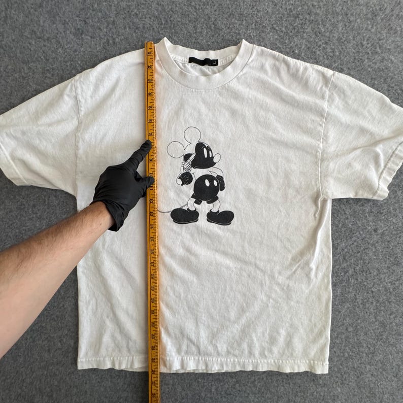 May include: White t-shirt featuring a black and white graphic of a cartoon mouse character. The shirt has a crew neck and short sleeves. A measuring tape is held up against the shirt.