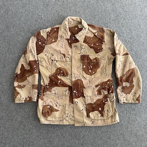May include: A desert camouflage jacket in tan and brown, featuring four front pockets and button fastenings. The long-sleeved jacket has a collared neckline and is displayed flat on a grey surface.