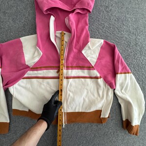 Op de afbeelding: Een cropped zip-up hoodie in roze, wit en bruin. De hoodie heeft een capuchon met trekkoord, een volledige rits en lange mouwen. Het ontwerp bevat horizontale bruine strepen over de borst en een bruine geribbelde zoom. De lengte wordt gemeten met een liniaal.
