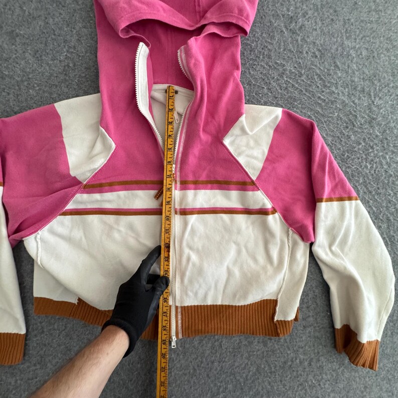 Op de afbeelding: Een cropped zip-up hoodie in roze, wit en bruin. De hoodie heeft een capuchon met trekkoord, een volledige rits en lange mouwen. Het ontwerp bevat horizontale bruine strepen over de borst en een bruine geribbelde zoom. De lengte wordt gemeten met een liniaal.
