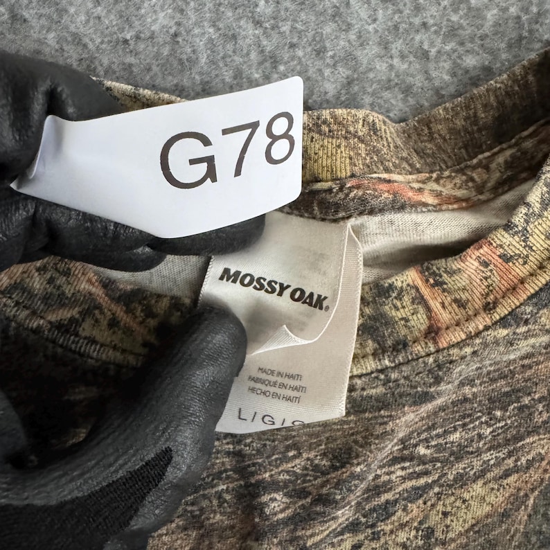 May include: A close-up of a camouflage-patterned shirt with the brand tag "Mossy Oak." The shirt is a blend of brown, green, and tan colors. A white tag with the text "G78" is visible. The shirt is a size L/G.