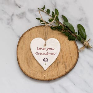 May include: A white heart-shaped ceramic ornament with the text "Love you Grandma" in burgundy, and a small flower illustration. The ornament hangs on a natural wood slice with a twine string, and is decorated with a sprig of greenery.