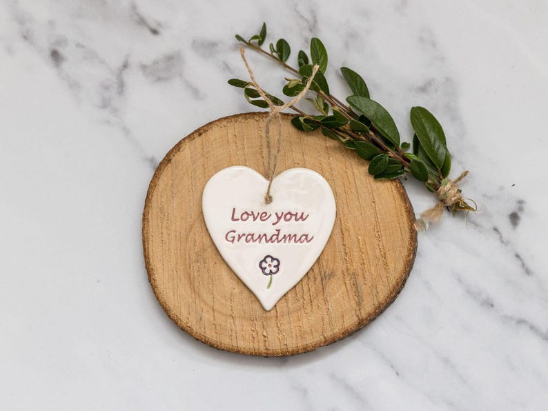 May include: A white heart-shaped ceramic ornament with the text "Love you Grandma" in burgundy, and a small flower illustration. The ornament hangs on a natural wood slice with a twine string, and is decorated with a sprig of greenery.