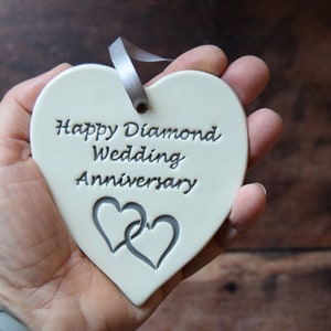 May include: A heart-shaped white ceramic ornament featuring the embossed text Happy Diamond Wedding Anniversary and two interlocking heart outlines. A grey ribbon is threaded through a hole at the top for hanging purposes.