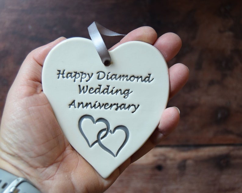 May include: A heart-shaped white ceramic ornament featuring the embossed text Happy Diamond Wedding Anniversary and two interlocking heart outlines. A grey ribbon is threaded through a hole at the top for hanging purposes.