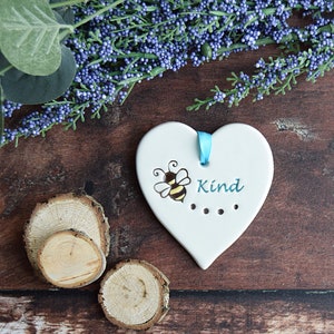May include: White ceramic heart-shaped ornament with a blue ribbon, a yellow and black bee illustration, and the word "Kind" in blue text.
