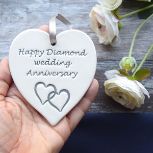 May include: A white heart-shaped ceramic ornament featuring the engraved text Happy Diamond wedding Anniversary and two interlocking heart outlines. A grey ribbon is threaded through a hole at the top for hanging purposes.