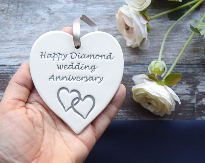 May include: A white heart-shaped ceramic ornament featuring the engraved text Happy Diamond wedding Anniversary and two interlocking heart outlines. A grey ribbon is threaded through a hole at the top for hanging purposes.