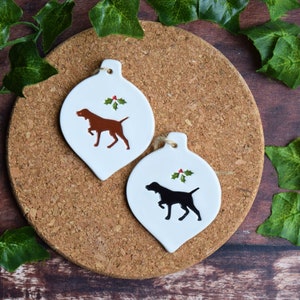 May include: Two white ceramic Christmas baubles shaped like teardrops with a brown and black dog silhouette on each. The baubles have a green holly sprig with red berries on each.