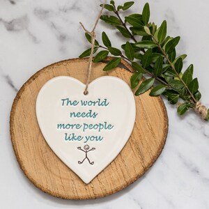 May include: White ceramic heart-shaped ornament with the text "The world needs more people like you" and a stick figure drawing. The ornament is on a wooden slice.