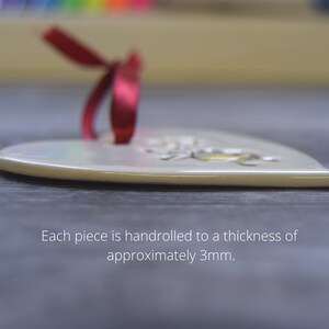 May include: A heart-shaped ceramic ornament with a smooth, off-white finish and a red ribbon loop attached at the top. The text on the image states: Each piece is handrolled to a thickness of approximately 3mm. The ornament has a flat profile.
