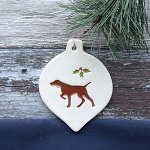 May include: White ceramic Christmas bauble shaped like a teardrop with a brown silhouette of a dog and a green holly sprig with red berries.