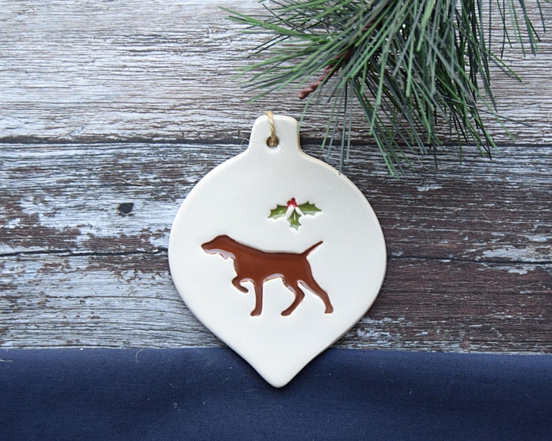 May include: White ceramic Christmas bauble shaped like a teardrop with a brown silhouette of a dog and a green holly sprig with red berries.