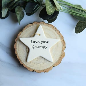 May include: A cream-coloured star-shaped ornament with the words "Love you Grumpy" in black. It sits on a wooden slice. Green foliage is visible in the background.